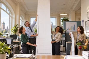 Three fictional civil servants laugh as they hold an absurdly tall blank graph in a bright statistics office while a shopper holds a grocery receipt.