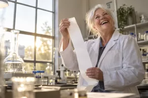 Original editorial photograph of a laughing older woman scientist alone in a sunlit laboratory holding a blank oversized receipt for UK research and development spending