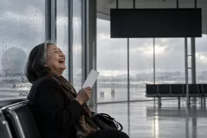 Original editorial photograph of a laughing older Asian woman alone beside an unbranded airport departure board with no text, holding a blank boarding card on a rainy day