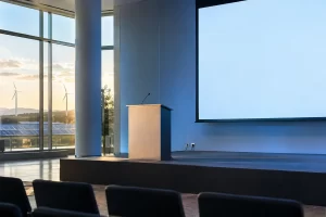 An empty modern conference stage with a blank projection screen and lectern looks out over wind turbines and solar panels.