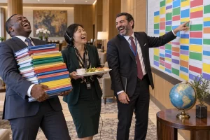 Three laughing delegates juggle colourful agendas, a lunch tray and a translation headset beside a crowded conference timetable.
