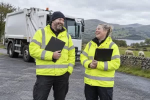 Two laughing environmental enforcement officers with blank clipboards beside a generic waste vehicle