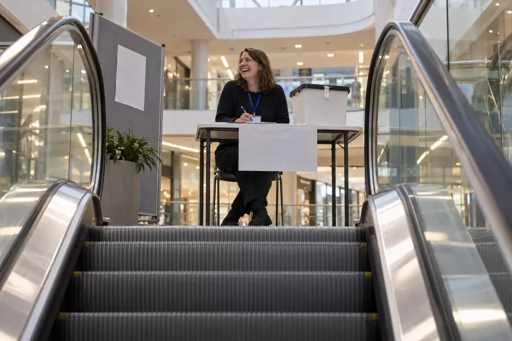 One laughing adult election worker sits at a generic ballot table near the top of a shopping-centre escalator, viewed from below.