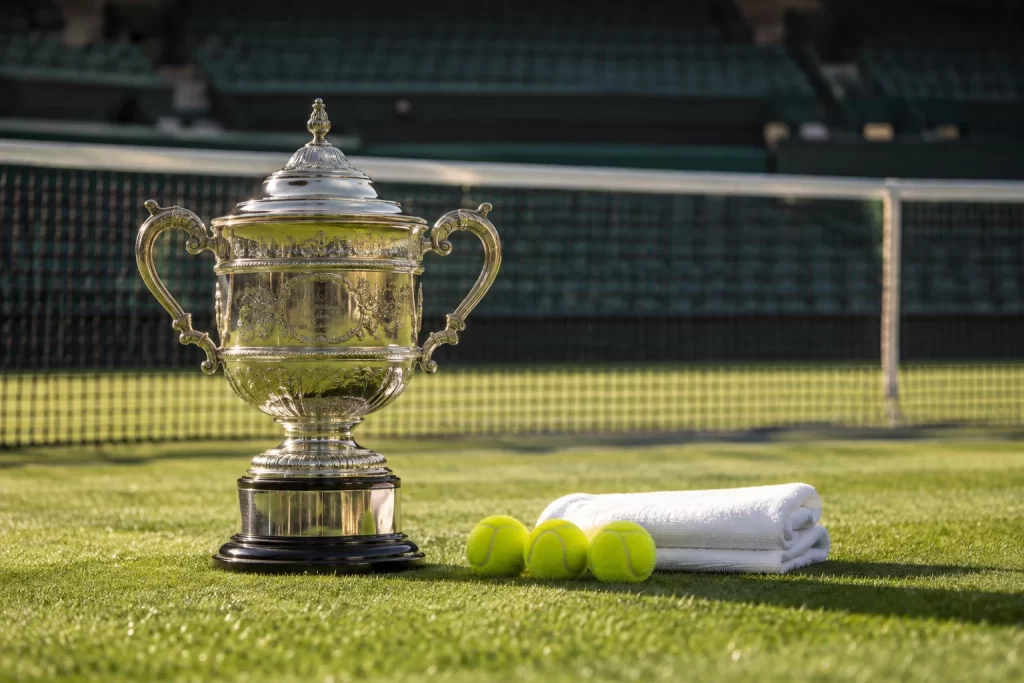 A silver tennis trophy, three tennis balls and folded towels rest beside the net on an empty grass court.