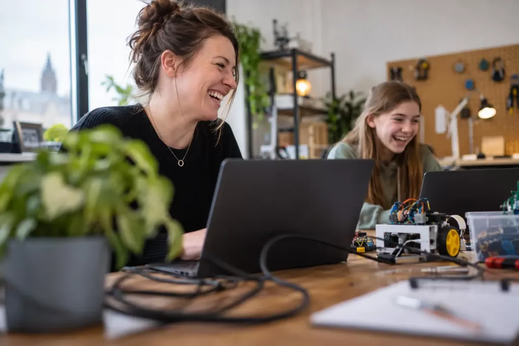 Laughing woman mentor and laughing teenage girl working with laptops and electronics in a British tech workshop