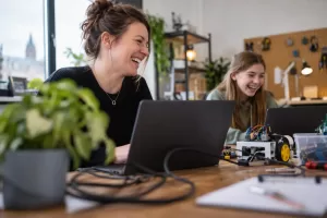 Laughing woman mentor and laughing teenage girl working with laptops and electronics in a British tech workshop