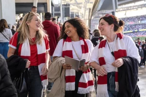 Three adult women football supporters laugh together on a bright stadium concourse before a cup final.