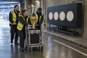 Three laughing stadium workers wheel a gold football trophy past a blank countdown display in a service tunnel.